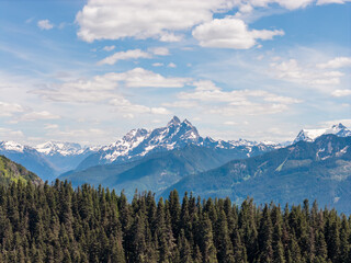 Fototapeta premium Snowcapped Mountain Range Above Evergreen Forest Under Blue Sky With Puffy Clouds Near Chilliwack, BC, Canada