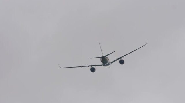 Commercial passenger jetliner flying away and climbing into grey overcast cloudy sky, telephoto low angle rear view
