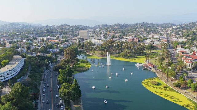 Scenic flight over Echo Park Lake in Los Angeles showing pedal boats floating on water with a large fountain, traffic moving on nearby road, urban landscape