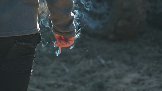 A man in a fleece jacket holds a cigarette in his lowered hand. Close-up. Slow motion of white smoke, outdoors