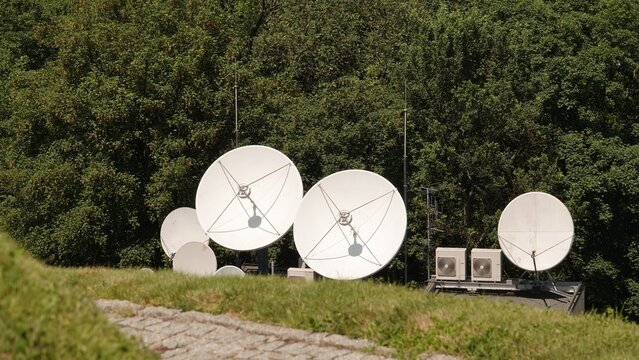 Grupa białych anten satelitarnych o r&oacute;żnych rozmiarach zainstalowana na dachu budynku obok urządzeń klimatyzacyjnych. W tle widoczna jest gęsta ściana zielonych drzew w słoneczny dzień.