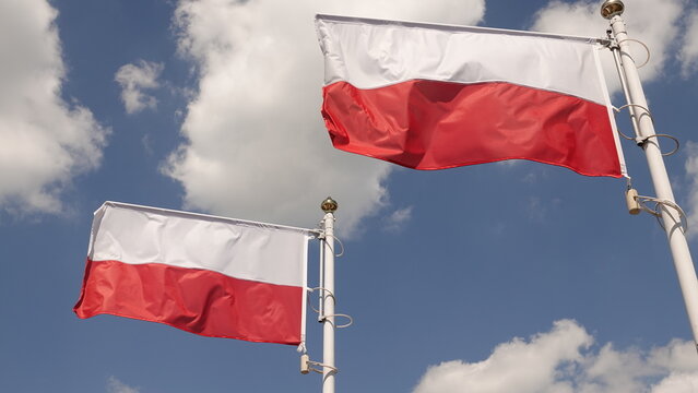 Dwie biało-czerwone flagi Polski powiewające na masztach na tle błękitnego nieba z białymi, puszystymi chmurami. Narodowe symbole uchwycone w pełnym słońcu symbolizują patriotyzm i wolność