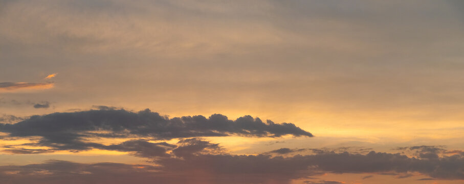 Panoramic sunset sky with warm golden-orange hues and a dark band of clouds on the horizon, romantic evening landscape