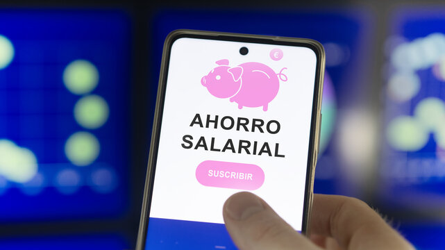 Close-up of a smartphone displaying a digital banking app with a piggy bank and Spanish text "Ahorro Salarial" Salary Savings. "Suscribir" Subscribe button, set against a blurred financial charts.