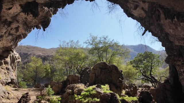 Natural cave entrance forming an arch, looking out towards a sunny, green landscape with trees and rugged hills under a clear blue sky in Dhofar, Oman, offering a sense of exploration