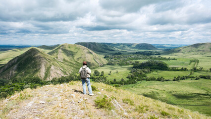 Fototapeta premium Alone backpacker woman stands on a rocky hillside overlooking a wide green valley with rolling mountains, scattered trees, and a cloudy sky in the background