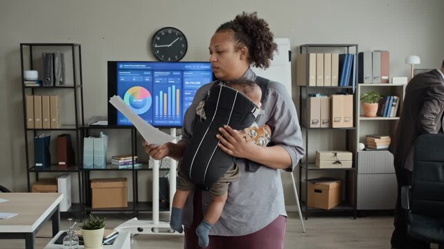 African American woman holding baby in carrier while reviewing documents in office. Video perfect for commercial use promoting work-life balance and diverse corporate culture
