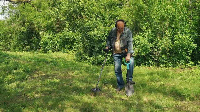 Treasure hunter in denim jacket and headphones operates a metal detector in a grassy area, bending down to examine the ground for buried items, surrounded by lush green foliage