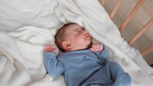 Baby boy sleeping with clenched fists and curled legs in a blue knit outfit on a white fleece blanket inside a wooden crib; gentle serenity.