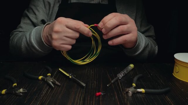 Hands of a male technician manipulate yellow and green electrical wires while preparing connectors with various tools on a wooden work surface