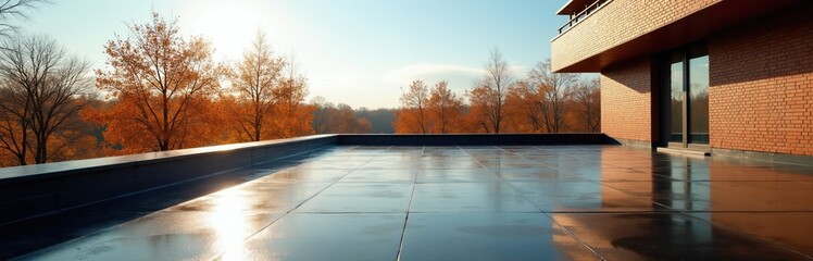 Obraz premium Modern rooftop terrace with slick dark tiles and brick building. Autumn trees with orange leaves behind railing. Sunny day, clear sky, peaceful landscape view.