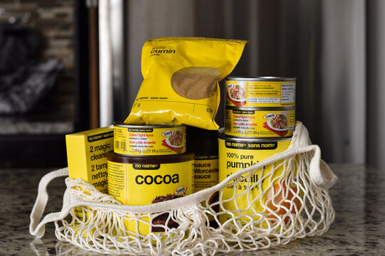 Assorted No Name budget friendly grocery products inside a reusable macrame mesh bag placed on a kitchen counter in a home interior setting