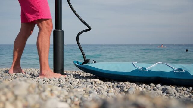 Close-up of a man's legs as he inflates a blue stand-up paddleboard with a hand pump on a pebble beach, preparing for a water sports activity on a sunny summer day by the sea