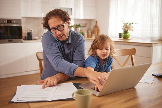 A father helps his child with a laptop while surrounded by documents on a table in their cozy kitchen. Sunlight streams through the window, creating a warm atmosphere.
