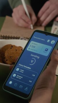 Hands of young student holding smartphone with mobile banking app interface while flatmate writes in notebook, calculating expenses. Fintech apps, digital payments, or budgeting, vertical
