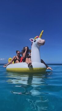 Two women and kids lounging on inflatable unicorn float in clear ocean, casual sunbathing and conversation, gentle reflection on water, relaxed social summer moment, bright sky and calm