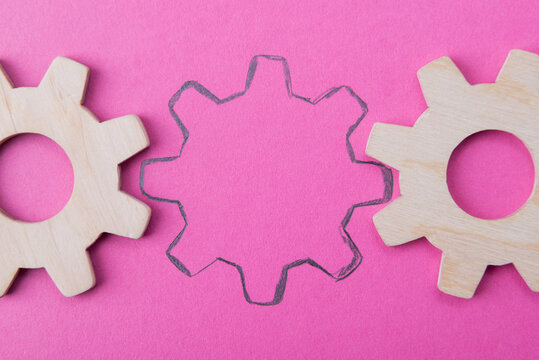 Missing gear wheel concept on a pink background representing a vacant position in business. Drawn outline of a cogwheel between wooden gears shows recruitment and hiring process.
