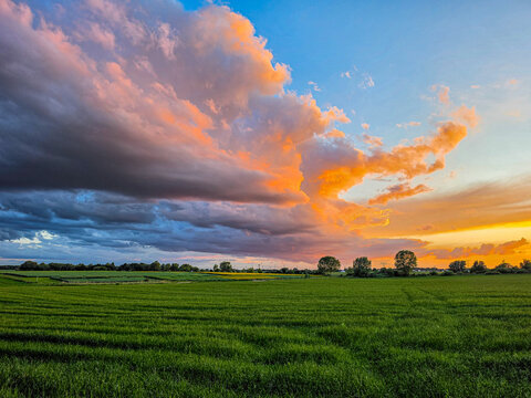 Vibrant rural landscape with lush green fields stretching toward the horizon beneath dramatic clouds lit by the setting sun. Strong color contrasts between the sky and farmland create a striking count