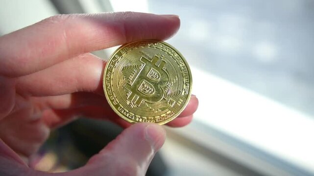 Human fingers holding shiny golden bitcoin coin against bright blurred background
