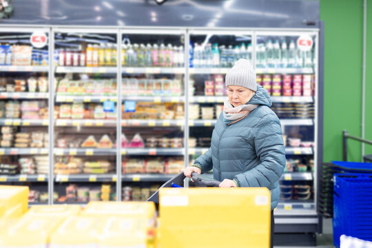 Elderly female customer wearing scarf choosing groceries while standing with trolley in contemporary retail store interior.