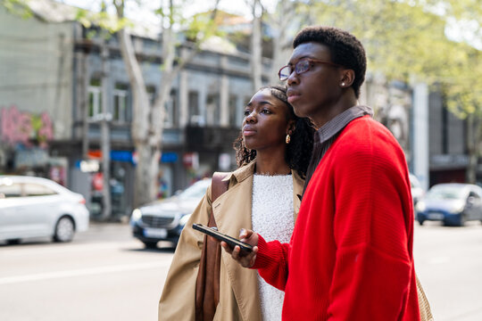Concentrated african american man and woman tourists waiting for taxi while standing on street, black male and female travelers friends ordered car online on phone and waiting for driver.