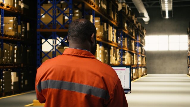African american male employee handling parcel logistics on laptop, distribution hub operations integrating shipment details and inventory management to support e-commerce efficiency.