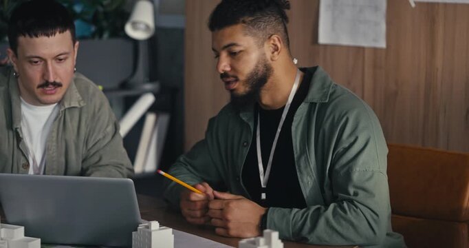 Two men sit at a table sharing ideas and looking at a laptop. They take notes and discuss their work in an office