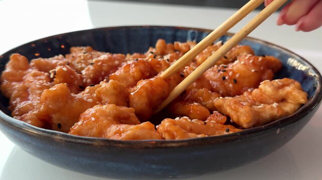 A person uses chopsticks to pick up and enjoy a chicken wing coated in sweet and sour sauce with sesame seeds
