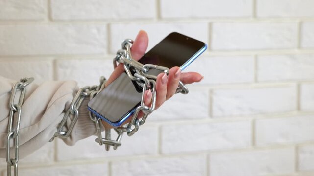 Woman chained to smartphone illustrating phone addiction concept. Woman's hand wrapped in a metal chain holding a mobile phone, symbolizing dependence on social media, internet slavery