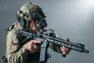 Obraz premium Male soldier in tactical gear aiming a rifle with a scope, wearing a helmet and night vision goggles, against a smoky gray background