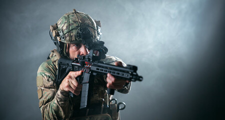 Obraz premium Male soldier in camouflage uniform aiming a rifle with a focused expression, smoke and dramatic lighting creating a tense atmosphere in a darkened environment