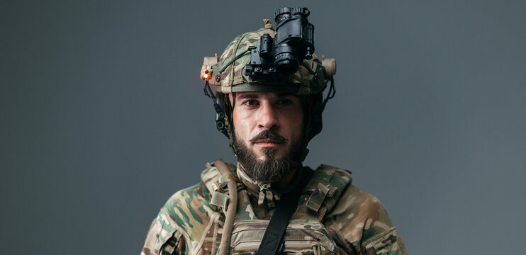 Male soldier wearing tactical gear and helmet with night vision device, posing against a gray background, showcasing military attire and equipment details