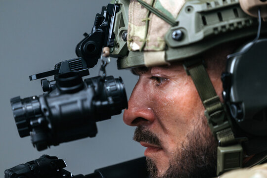 Armed special forces operator in tactical gear with weapon raised, equipped with night vision device, focused expression, and camouflage helmet against a neutral background