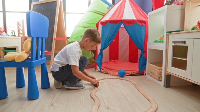 Boy kneeling on the floor pushing a wooden toy train along a curved track beside a striped play tent and blue chair in a bright playroom; concentration imaginative play.