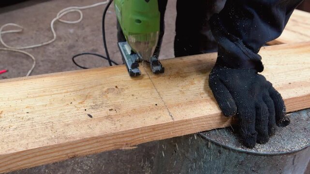 Close-up of a carpenter wearing protective gloves cutting a wooden plank with a green electric jigsaw, creating a cloud of sawdust in a workshop environment during a home improvement project