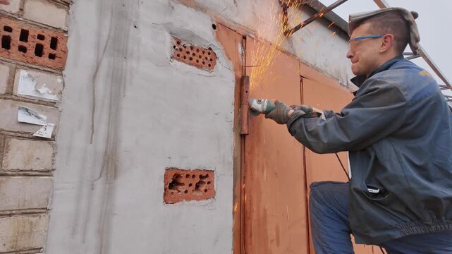 Professional male worker in protective glasses and gloves using an angle grinder to cut through a rusty metal door hinge, creating a shower of bright orange sparks against the brick wall