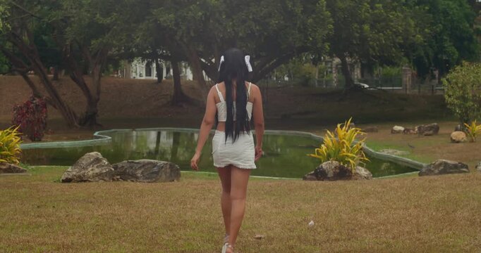 Creamy bokeh background of park trees as a light-skinned girl in a white mini skirt walks into focus. Soft, overcast daylight.