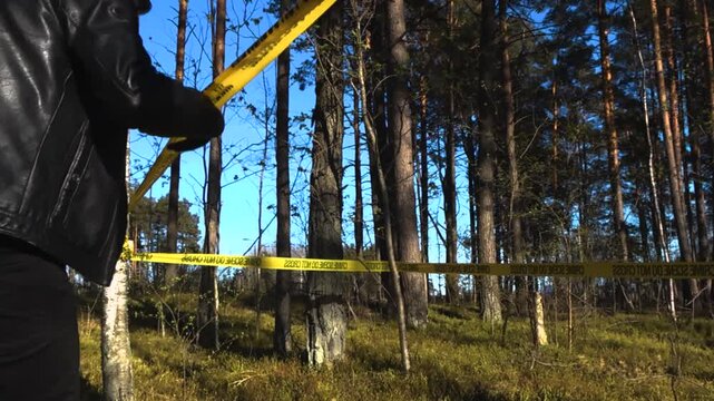 Undercover police officer or detective in a black leather jacket carefully crossing under the yellow barrier tape at a crime scene in a forest