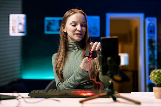 Joyous Gen Z entertainer presenting wired gaming mouse in sponsored video, filming with mobile phone, with upbeat tech guru promotes computer peripherals, recording with smartphone on tripod.