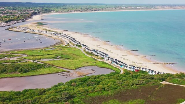 Drone footage looking across Mudeford Sandbank with beach huts sandy beach turquoise sea Christchurch Harbour wetlands boats and coastal landscape.