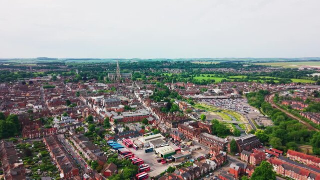 Wide drone footage of Salisbury city centre in Wiltshire, England, showing Salisbury Cathedral, residential streets, car parks, railway lines, roads, green spaces and surrounding countryside.