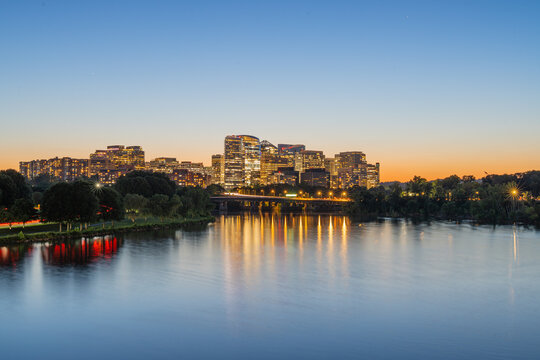 Arlington, Virginia, long-exposure photo of the illuminated Rosslyn skyline reflecting perfectly across the calm surface of the Potomac River during a vibrant twilight sunset.