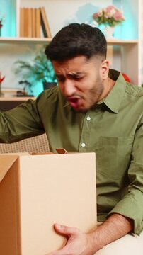 Indian man on home sofa opens delivery box and frowns at wrong order with damaged goods. Arabian boy on home sofa spreads hands in anger and reacts to online shopping problem with consumer complaint