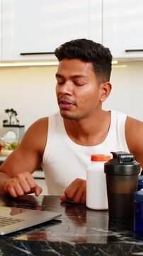 Indian man explains sports nutrition and workout supplements on laptop during meal planning at home. Arabian boy shares training advice and protein diet tips with focused face in home kitchen indoors