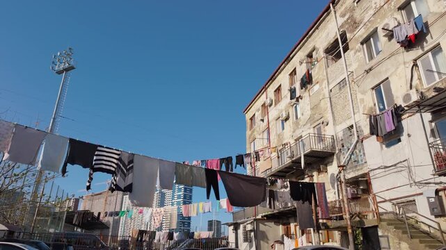 Low-angle view of laundry drying on clotheslines strung between old, weathered apartment buildings in a poor neighborhood, showing a stark contrast with modern skyscrapers in the distance