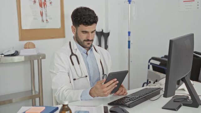 Young hispanic doctor in a clinic holds a tablet and scrolls with fingers, stethoscope visible at desk with monitor and anatomy models; calm professionalism.