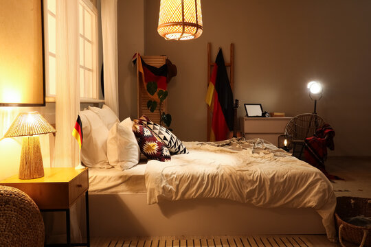 Interior of stylish bedroom with cozy bed and German flag hanging on folding screen at evening