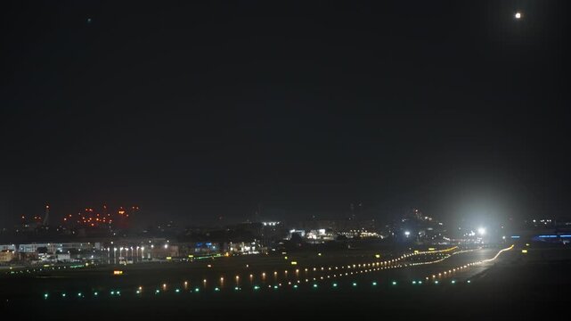 Wide view of airport runway and taxiway lights glowing at night, green and white lights in rows, city skyline with red beacons and bright moon in the dark sky