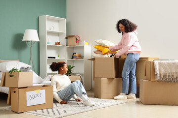 Happy African-American woman and her daughter packing lamp into cardboard boxes in bedroom on...