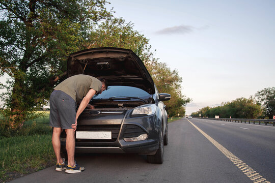 Man examining engine under open hood of broken car parked on highway shoulder, roadside vehicle failure during journey, transportation problem concept, automotive diagnostics, emergency 